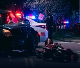 A police officer stands beside a severely damaged motorcycle lying on its side next to a dented dark SUV at a nighttime accident scene with flashing emergency lights.